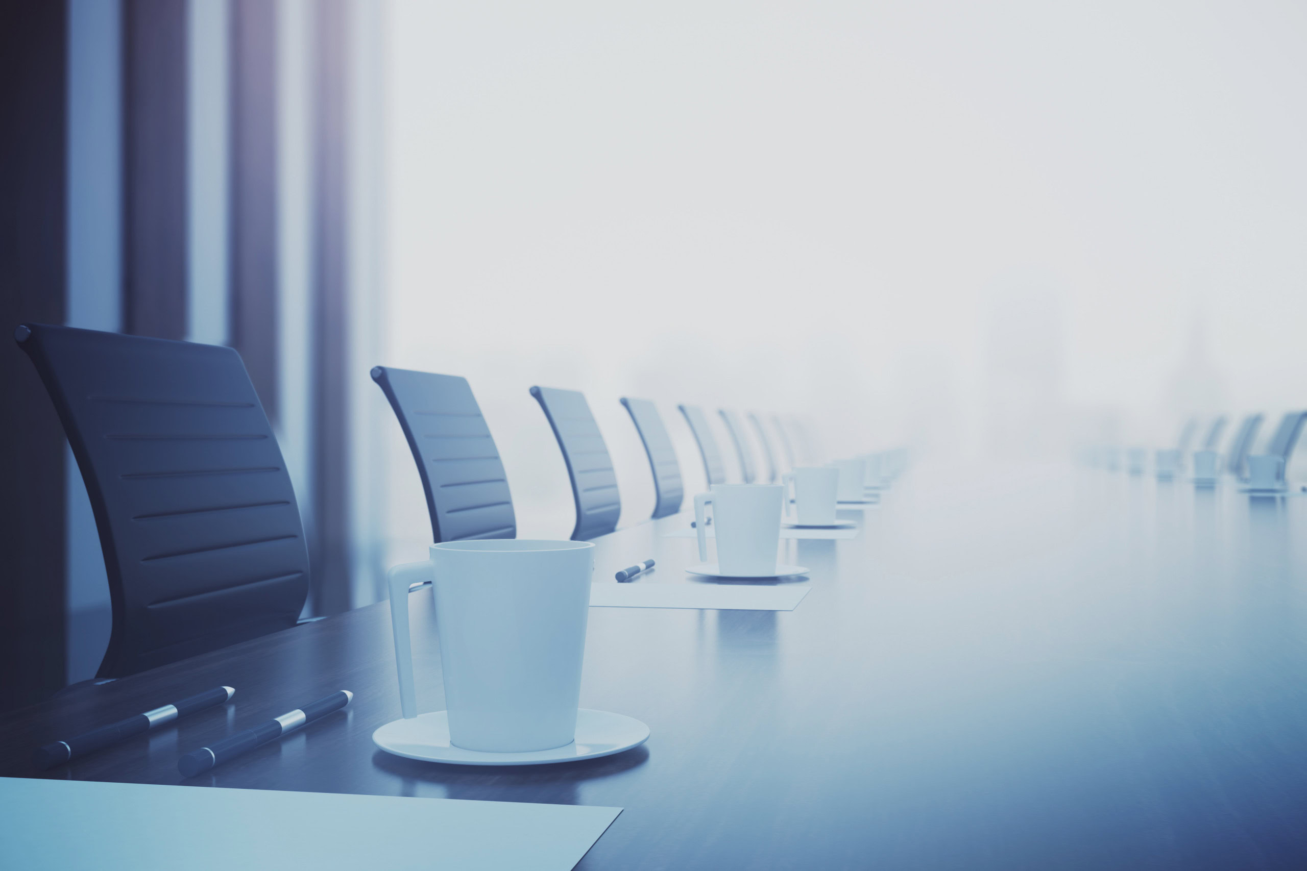 A long conference table with several empty black chairs, white coffee cups on saucers, and a few notepads with pens.