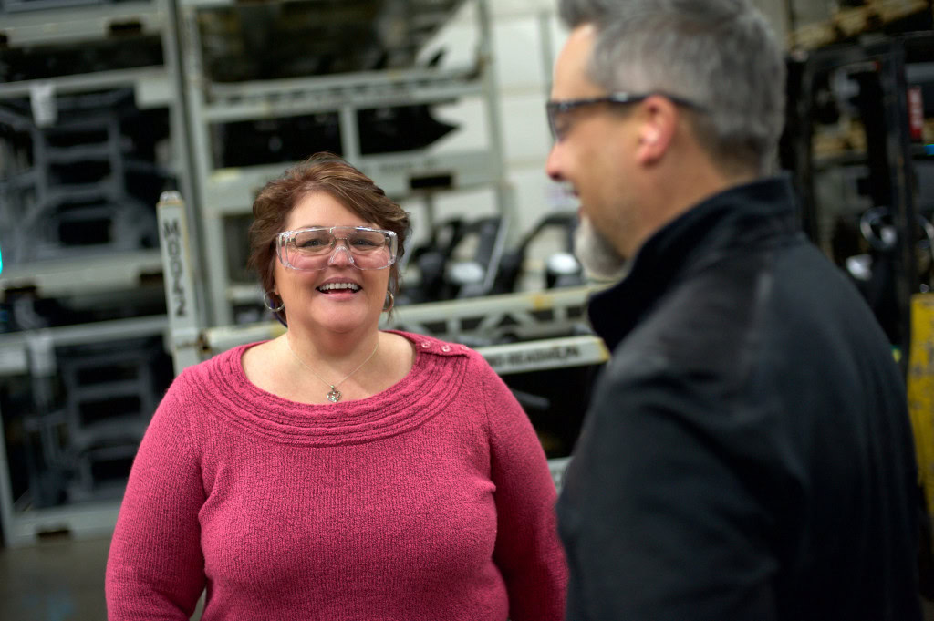 A woman wearing a pink sweater and safety glasses is smiling while talking to a man in a black jacket and safety glasses.