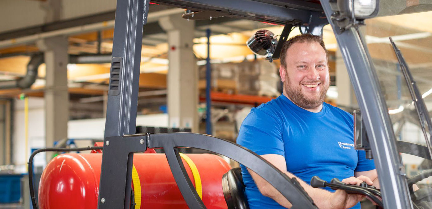 Un hombre con camisa azul sonríe sentado en el asiento del conductor'de una carretilla elevadora dentro de un almacén. La carretilla elevadora, con una cisterna roja adosada a la parte trasera, navega entre hileras de estanterías industriales y equipos