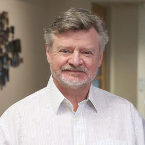 Peter Rye, a man with short gray hair and a beard is smiling at the camera. He is wearing a striped white dress shirt and standing indoors with a blurred background.