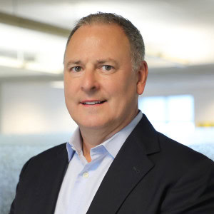 A middle-aged man with short, gray hair is wearing a dark blazer over a light-colored shirt. Tim Joseph is smiling and standing in a bright office environment with blurred background elements.