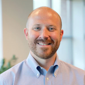 Walter Banta, a man with a beard and a shaved head is smiling at the camera. He is wearing a light blue and white checkered button-up shirt. The background is an indoor setting with blurred elements.